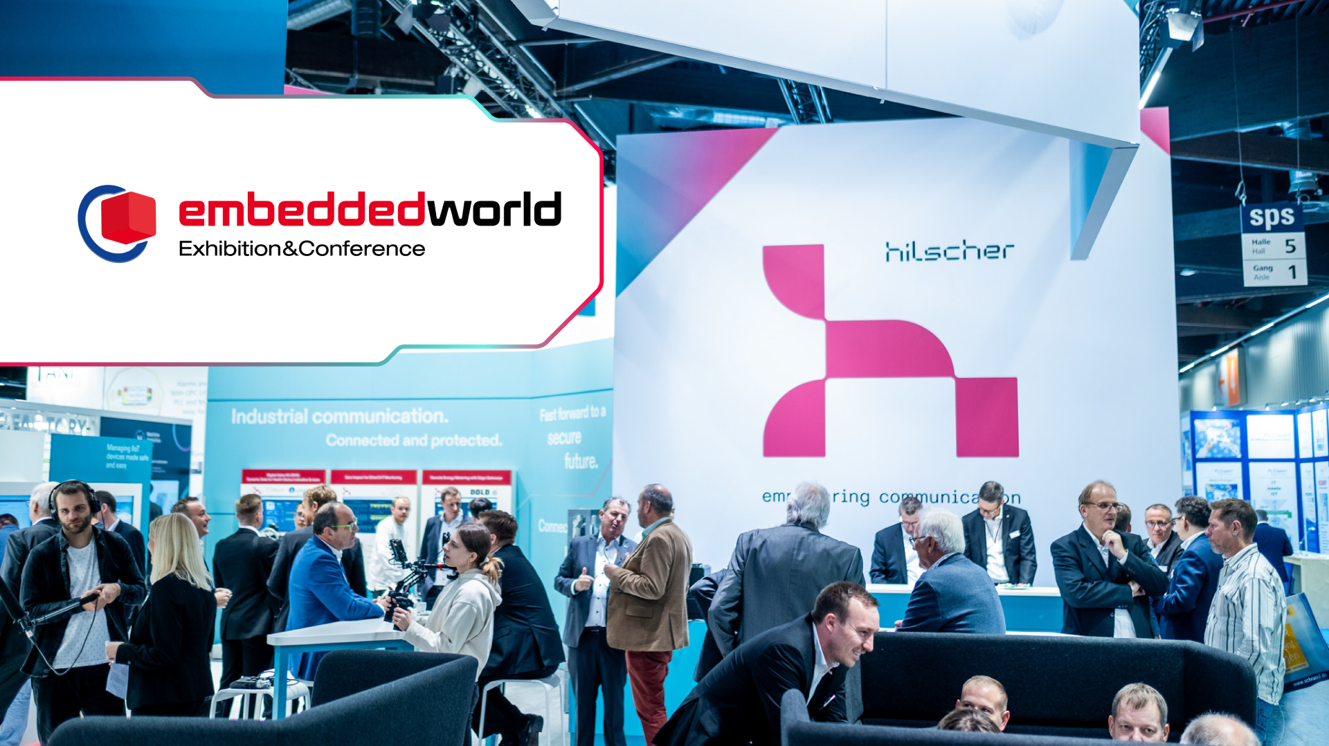 A crowded trade fair booth with numerous people in deep discussion. In the back is a large Hilscher logo on white background. In the top right corner is the embedded world logo.